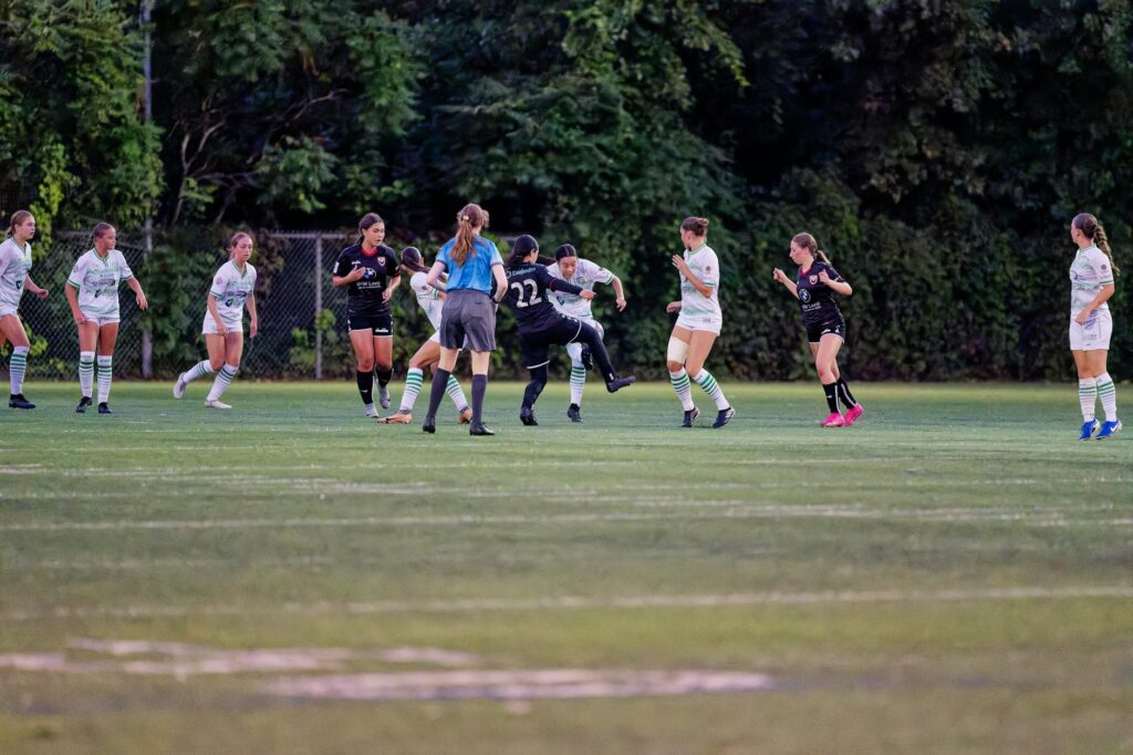 Female soccer match: players in white and dark kits chase the ball on a green field with a referee nearby.